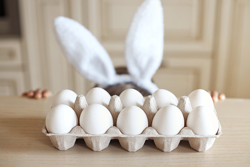 No face cute child with bunny ears is hiding under the table. White organic chicken eggs in a craft box stand on the table in the kitchen. Easter concept.