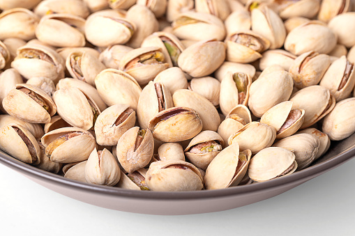 Pistachios in their shells, roasted and salted, in a brown bowl. Crunchy roasted fruits of Pistacia vera, with opened shells and seeds within. Snack food. Close-up, front view, macro food photo.