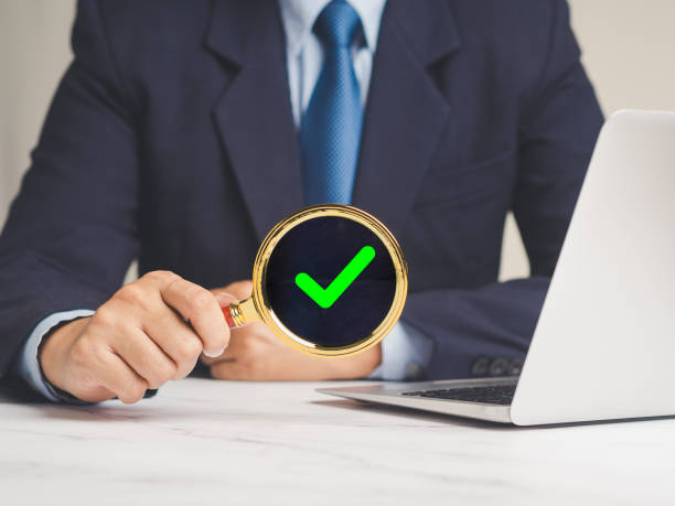 istock Businessman holding a magnifying glass with a green check mark symbol while sitting at the table 1906428775
