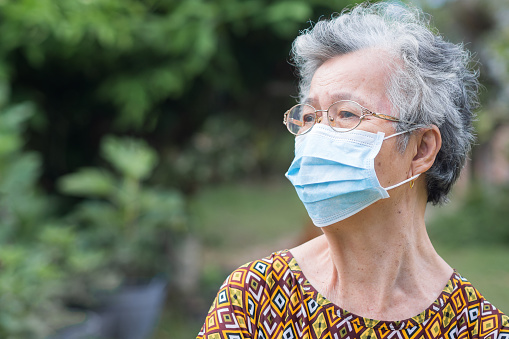 Senior woman wearing a surgical mask and looking away while standing in a garden. Mask for protect virus, coronavirus, pollen grains. Concept of old people and healthcare.