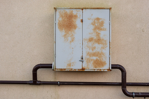 Gray metal box for gas equipment on a yellow wall of the house. There are brown curved pipes. Dots of rust are visible on the surface of the doors. Background.
