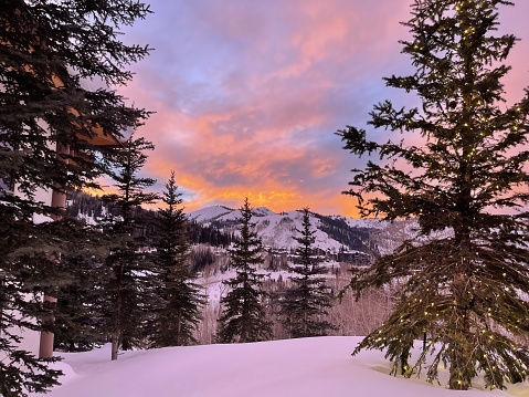 This photograph depicts a stunning sunset with a vibrant sky over a snowy mountain landscape, framed by evergreen trees in Deer Valley, Utah.