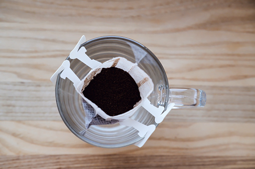 drip coffee with ground black Arabica coffee in a large glass cup close-up. portion of drip coffee per cup. a cup with paper drip coffee stands on a wooden board on a white background close-up.