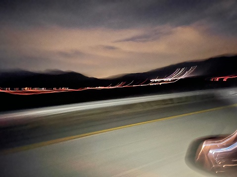 This photograph depicts a motion-blurred nighttime drive down a highway, with streaks of light indicating movement and a mountainous silhouette in the distance.