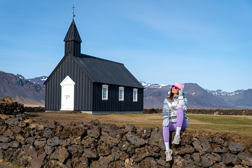 A young woman wearing a bright yellow raincoat poses atop a stone wall in front of a rural church