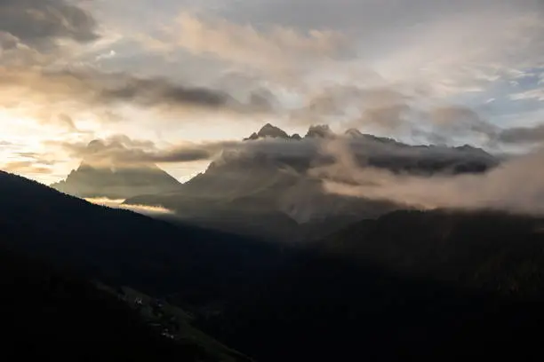 A picture of clouds and fog surrounding the Peitlerkofel mountain range. The sun is rising behind and to the left, shining rays of light down the valley.