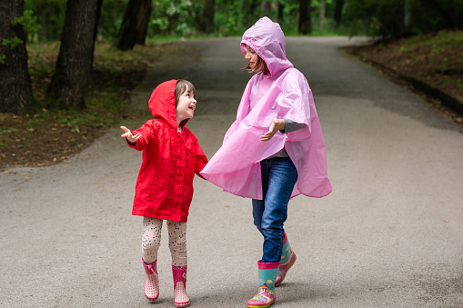 Two happy girls are walking in the park with raincoats on a spring day. They are holding hands and talking.