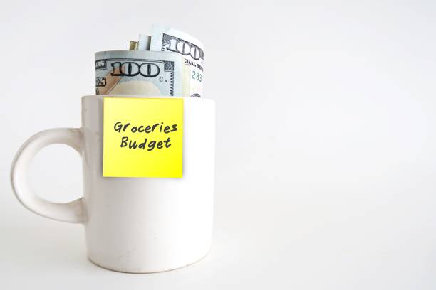 istock A white ceramic cup with cash dollars money inside, with yellow paper stick note written GROCERIES BUDGET, isolated on white background. Concept of setting limited budget for groceries shopping spending 1906662407