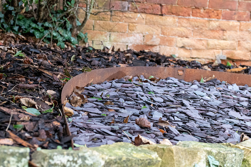 Slate pieces enclosed by a copper curved edging strip with bark chippings on one side and a weathered stone wall on the other against an aging brick wall