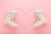 Heart shape created from white shoelaces between new beige warm winter boots on light pink table background. Pastel color. Female footwear. Closeup. Top down view.