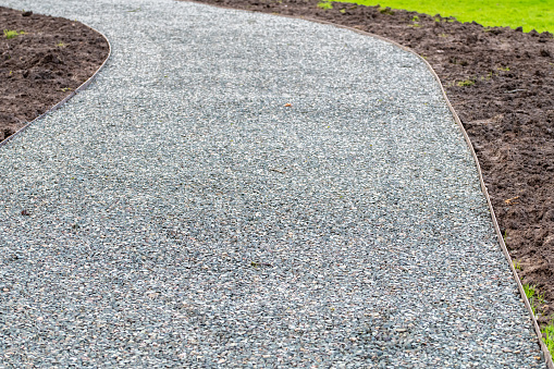 A curved corner section of a gray grey gravel driveway, footpath with freshly laid soil mud over green grass with clean steel edging strips