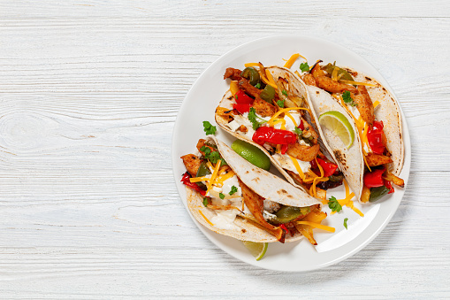 baked tex-mex chicken fajitas with mixed sweet pepper, onion, sour cream, cheddar cheese, lime, parsley and gluten free corn tortillas on plate on white wooden table, free space, flat lay