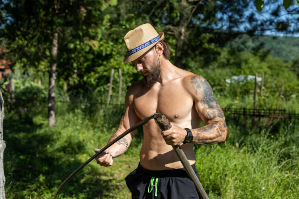A good-looking man is scything the grass shirtless.