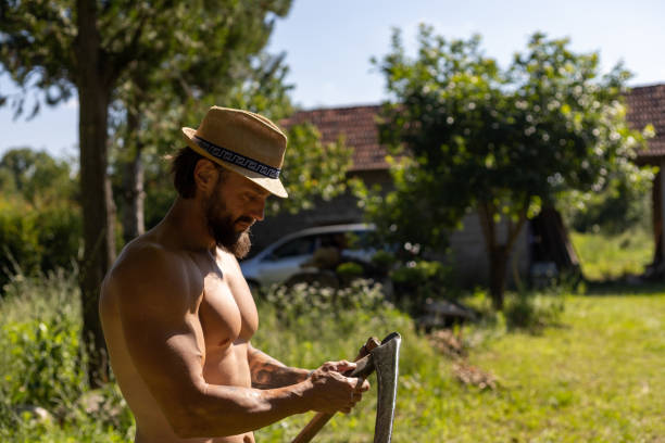 A good-looking man is scything the grass shirtless.
