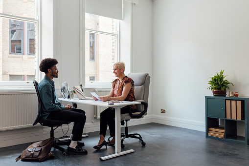 Side wide full length view of a mature female manager engaging in a focused discussion with a young male employee at the managers desk in an office interior in Newcastle, England. They are wearing smart casual clothing.

Videos also available for this scenario.