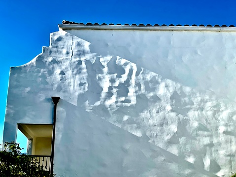 This photograph features a white building with a white wall and a white roof timed precisely to cast a magnificent section of light on the uneven plaster of a wall.