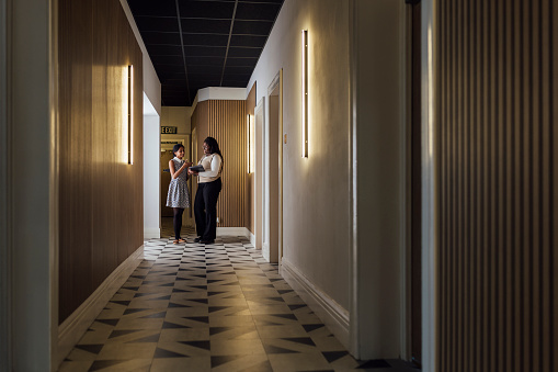 Full length wide front view of two young businesswomen standing in an office corridor in Newcastle, England. They are using a laptop to work as a team to complete a work task.