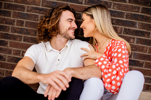Handsome ymiling young couple in love sitting in front of house brick wall