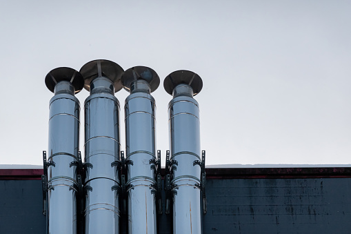 Four steel chimneys on the roof of the building. The pipes have a silvery surface. There is a thin layer of snow on the roof. Background.
