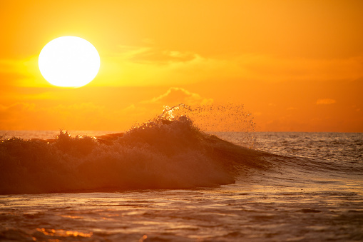 A breathtaking view of the sun setting over the ocean, casting an orange glow across the sky and water. The sun is partially obscured by distant clouds, creating a dramatic effect with rays of light piercing through. The ocean waves are highlighted by the sunlight, showcasing their texture and movement. The horizon is visible, separating the ocean from the sky and emphasizing the vastness of both elements. There’s a serene yet dynamic mood created by the combination of calm skies and active waters.