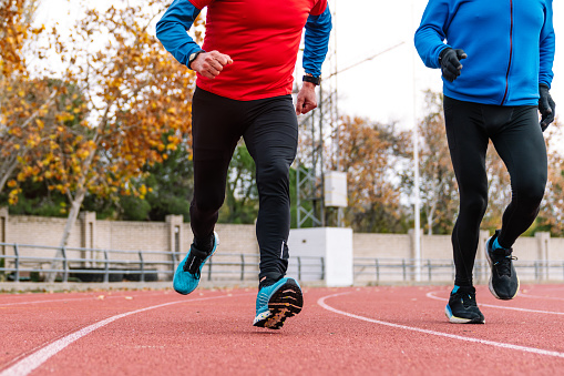 Two senior male runners in sportswear jogging on a red race track surrounded by autumn trees, displaying vitality and endurance.