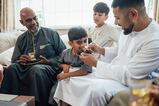 A shot of a group of family members sitting together on a sofa, talking and smiling as one of the males offers a child a drink from a cup of tea. They are gathered together for Eid celebrations at home in Middlesbrough, North East England. They are all dressed in traditional outfits for the occasion.