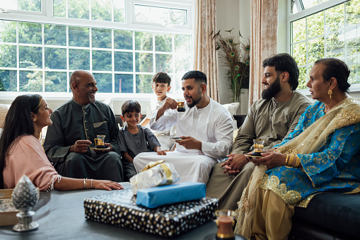 A shot of a group of family members sitting together on a sofa, talking and smiling as they enjoy cups of tea. They are gathered together for Eid celebrations at home in Middlesbrough, North East England. They are all dressed in traditional outfits for the occasion.