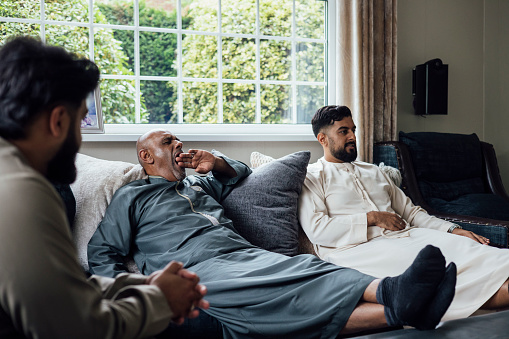 A shot of a group of family members taking a break and sitting together on a sofa with their feet up on the coffee table. They are gathered together for Eid celebrations at home in Middlesbrough, North East England. They are all dressed in traditional outfits for the occasion, one of them is yawning with one hand raised to cover his mouth.