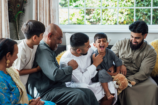 A shot of a group of family members sitting together on a sofa, laughing and having fun. There are five adults and two children. They are gathered together for Eid celebrations at home in Middlesbrough, North East England. They are all looking at one young boy who is being tickled, and they are dressed in traditional outfits for the occasion.