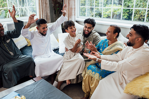 A shot of a group of family members sitting together on a sofa, laughing and having fun. They are gathered together for Eid celebrations at home in Middlesbrough, North East England. They are all looking at one young boy who is holding a small ball, and they are dressed in traditional outfits for the occasion.