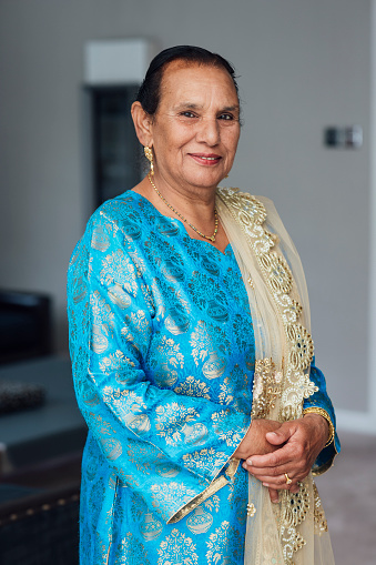 A three-quarter-length portrait of a senior adult female standing with her hands clasped in front of her in a home in Middlesbrough, North East England. She is smiling and dressed in elegant traditional clothing to celebrate Eid with family.