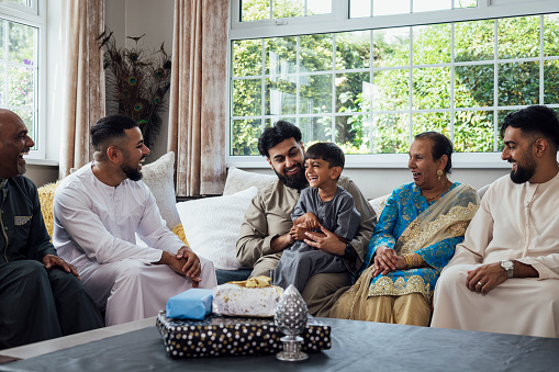 A shot of a group of family members sitting together on a sofa, talking and smiling. A young boy sits on the knee of an adult male and is smiling broadly. They are gathered together for Eid celebrations at home in Middlesbrough, North East England. They are all dressed in traditional outfits for the occasion.