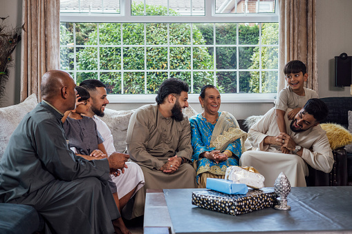 A shot of a group of family members sitting together on a sofa, talking and smiling. A young boy embraces an adult male from behind and they laugh. They are gathered together for Eid celebrations at home in Middlesbrough, North East England. They are all dressed in traditional outfits for the occasion.