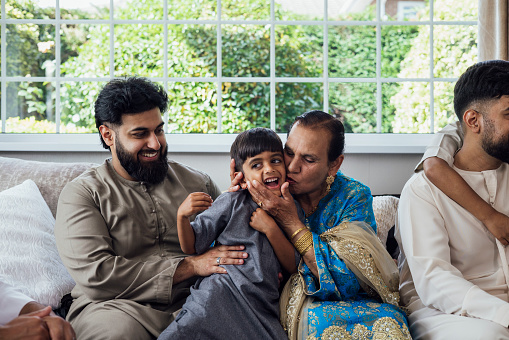A shot of a group of family members sitting together on a sofa, the grandmother kissing her grandson on the cheek, cupping his head between her hands as he squirms and smiles broadly. They are gathered together for Eid celebrations at home in Middlesbrough, North East England. They are all dressed in traditional outfits for the occasion.