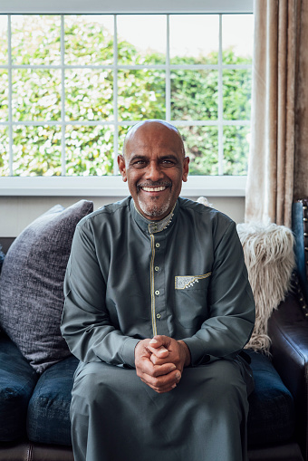A three-quarter-length portrait of a mature adult male sitting on a sofa with his hands clasped in front of him at home in Middlesbrough, North East England. He is smiling and dressed in elegant traditional clothing to celebrate Eid with family.