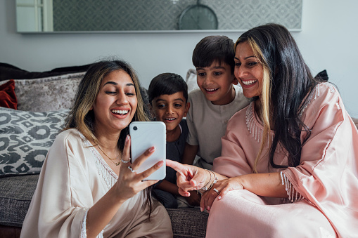 A shot of two adult females and two young boys sitting together in a living room at home in Middlesbrough, North East England. They are smiling whilst looking at selfies they have just taken on a mobile phone. One o the females points to the screen. They are gathered with family for Eid celebrations.