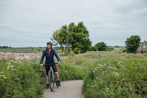 A mature woman wearing mountain biking sports clothing on an overcast day in Alnmouth, Northumberland, Northeastern England. She is on a bike ride along a bicycle trail that follows the coastline.