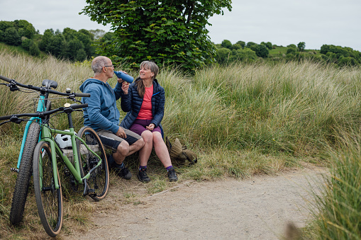 A mature, married couple wearing mountain biking sports clothing on an overcast day in Alnmouth, Northumberland, Northeastern England. They are on a bike ride along a bicycle trail that follows the coastline. They have stopped for a rest, a chat and a drink on a bench.