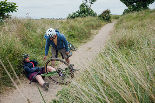 A mature, married couple wearing mountain biking sports clothing on an overcast day in Alnmouth, Northumberland, Northeastern England. The woman has fallen off her bike and is in pain as her husband tends to her.