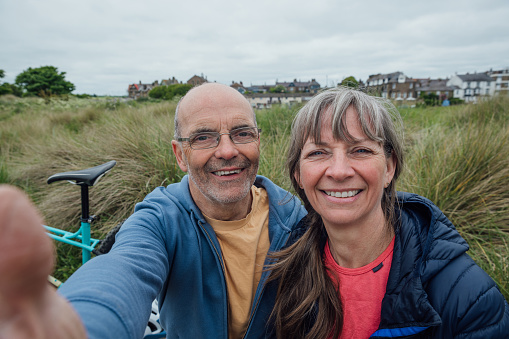 A mature, married couple wearing mountain biking sports clothing on an overcast day in Alnmouth, Northumberland, Northeastern England. They are on a bike ride along a bicycle trail that follows the coastline. They have stopped for a rest on a bench and are taking a selfie together on a smartphone.