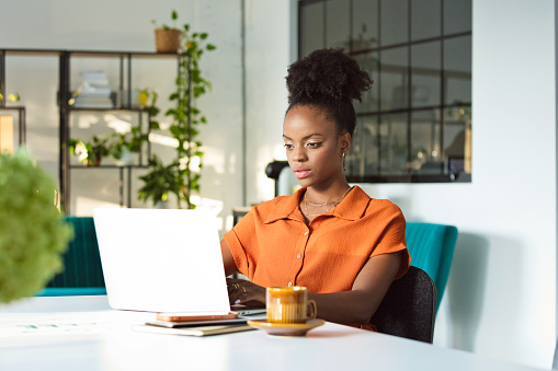 Beautiful, black women wearing orange dress sitting at the table in an office and working on laptop.