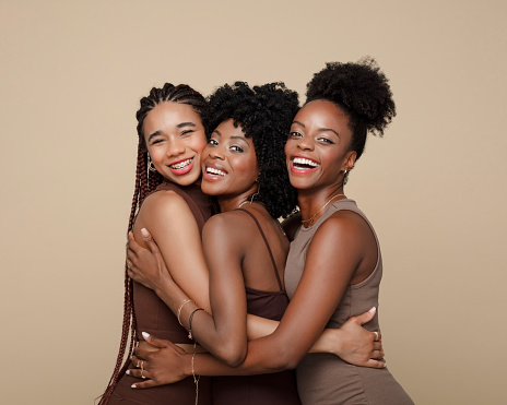 Three beautiful young women wearing brown clothes standing against beige background, embracing and smiling at camera.