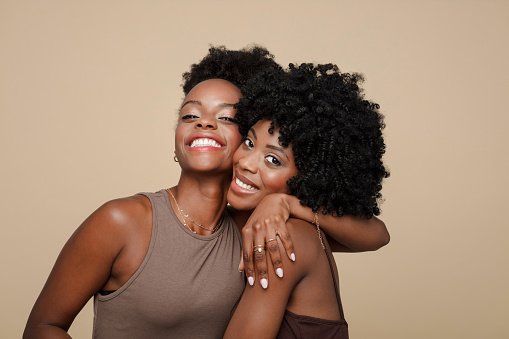 Two beautiful young women wearing brown clothes standing against beige background, embracing and smiling at camera.