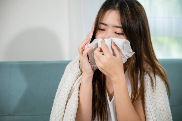 istock woman sick sitting down on sofa with cold blowing her nose hold tissues and talking on cellphone to doctor 1907002800