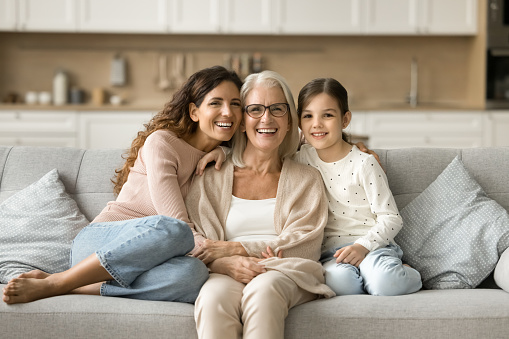 Happy grandma, mom and granddaughter kid sitting close on home sofa. Young mother and girl hugging grandmother with love, affection, laughing, smiling. Three female generations portrait
