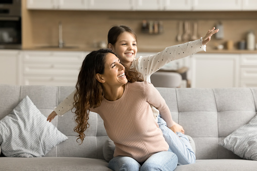 Positive playful pretty mom piggybacking little tween daughter with open arms. Sweet girl playing flying airplane on mothers back, enjoying activity on home sofa