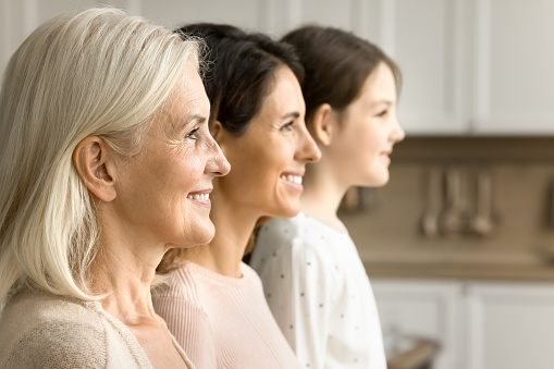 Cheerful blonde elderly grandmother looking forward away side shot with brunette daughter woman and grandkid girl standing in line in blurred background. Three female generations family portrait