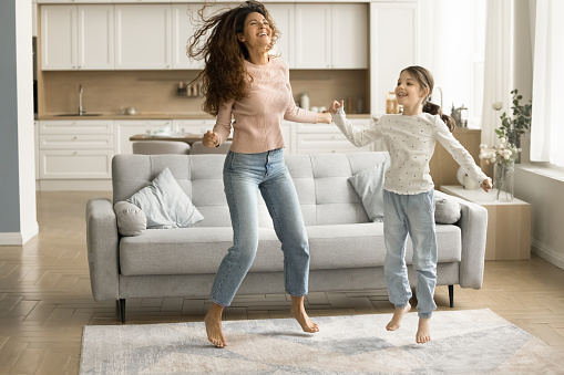 Cheerful active kid girl and excited mom hopping, jumping to music in living room, dancing together, enjoying home activity, family leisure, having fun, laughing, singing song. Full length portrait