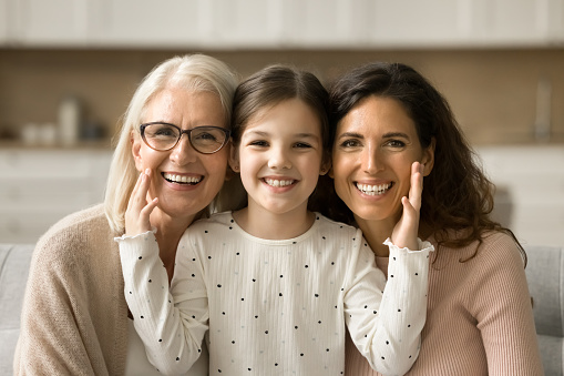 Cheerful cute little girl, mom, grandmother sitting close indoors, hugging, looking at camera with toothy smiles. Happy daughter kid touching cheeks of younger mother and grandma head shot portrait
