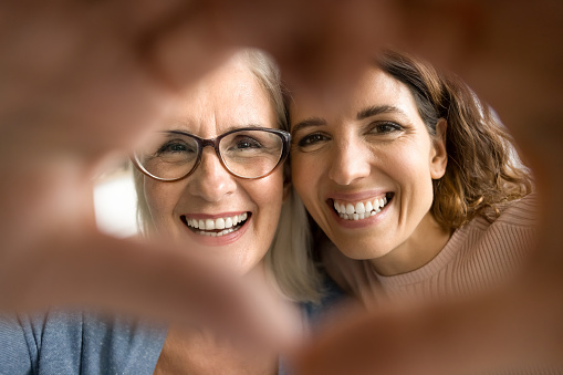 Cheerful pretty senior mom and adult daughter looking at camera through hand heart, making finger frame, smiling with white healthy teeth, expressing love, family affection, care. Close up portrait